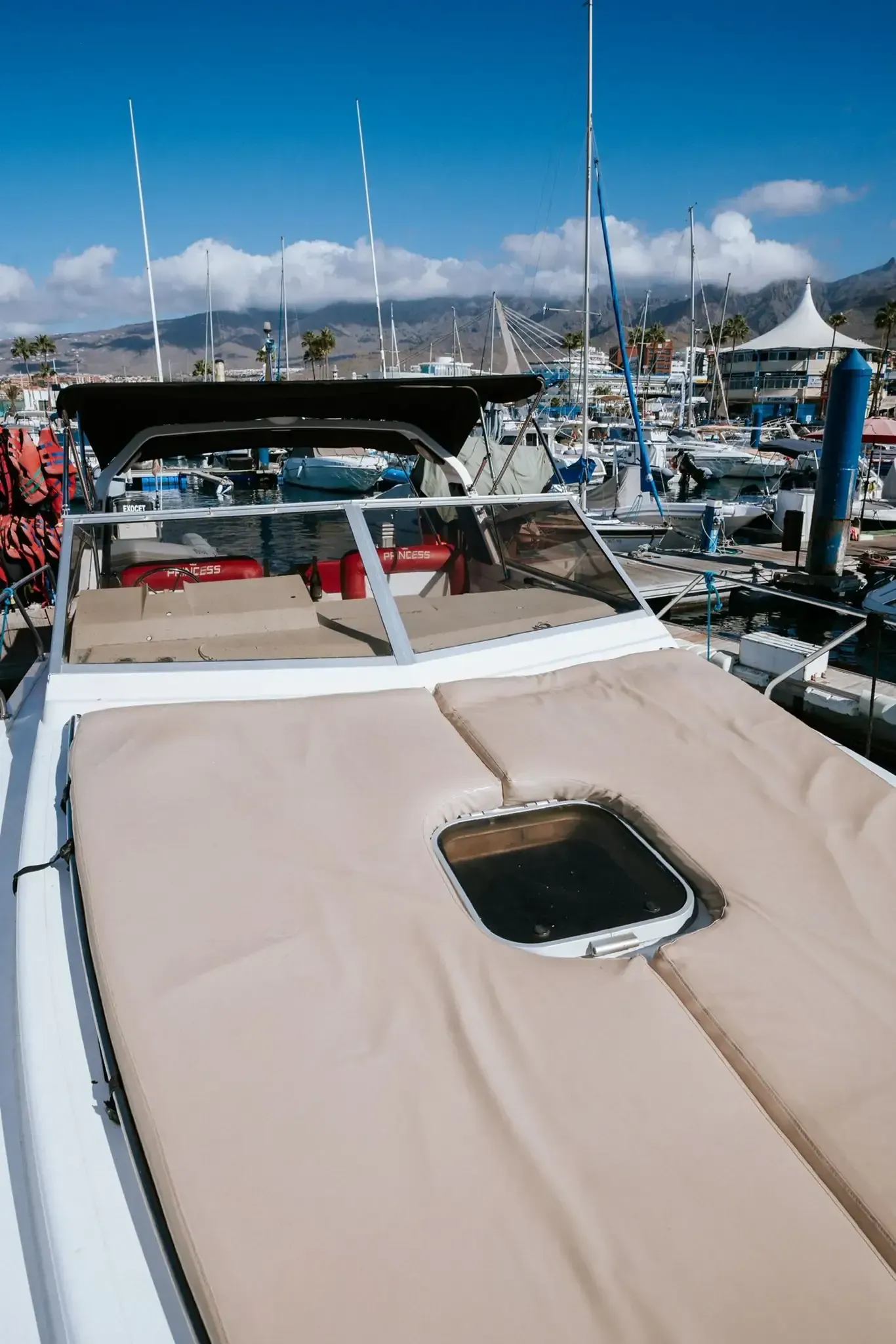 Un barco blanco con cojines beige y una pequeña ventana, atracado en un puerto deportivo en un día soleado.