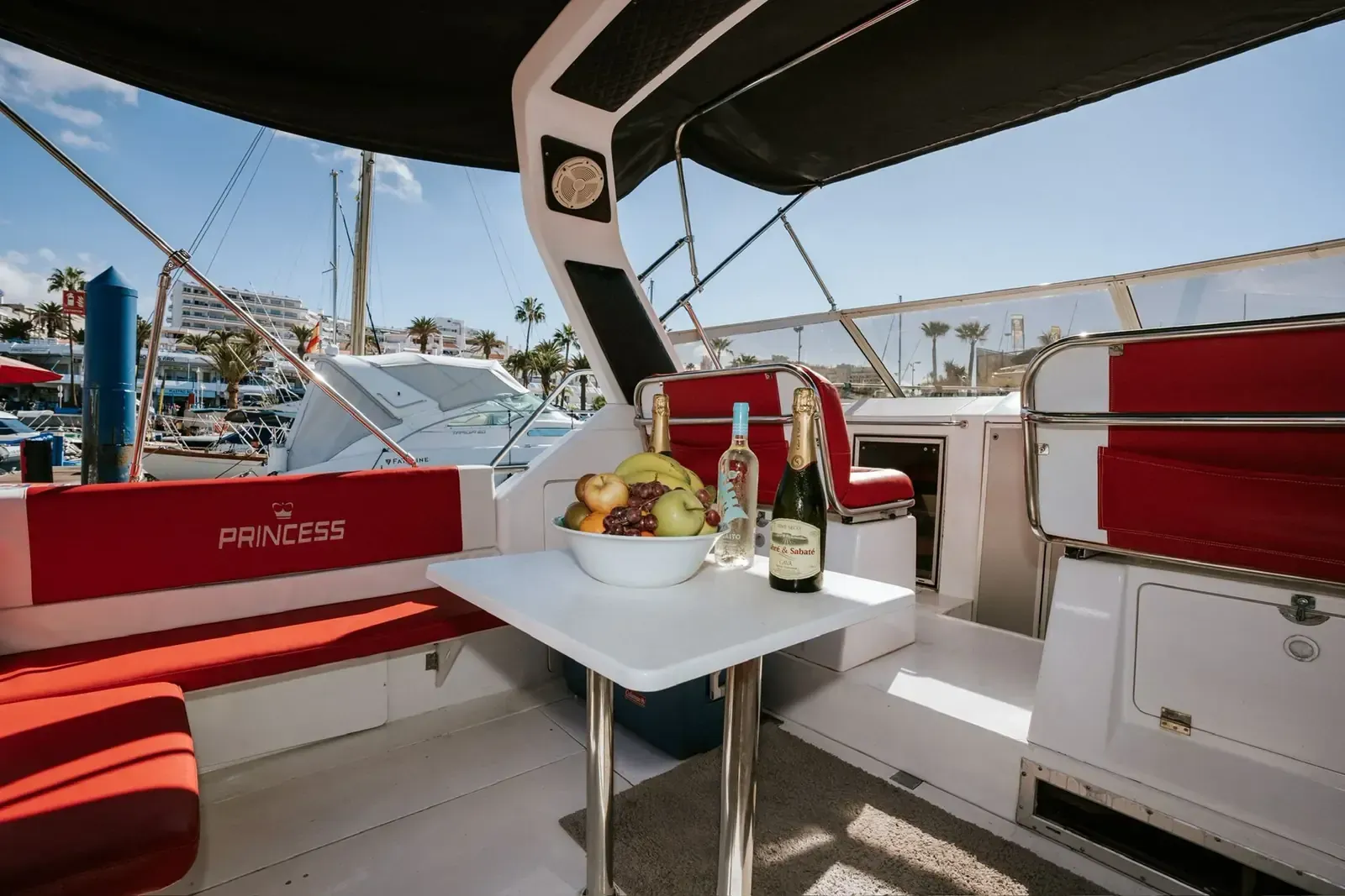 Interior del barco con asientos rojos, mesa con frutas y bebidas, vista soleada del puerto.