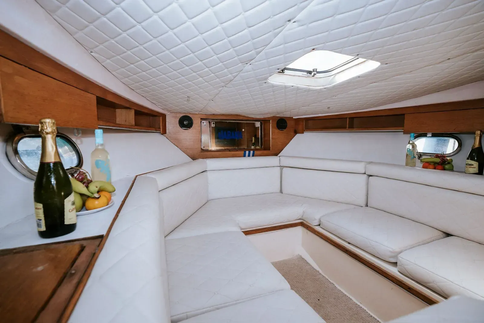 Interior de un yate con asientos de cuero blanco, paneles de madera y un pequeño tragaluz.