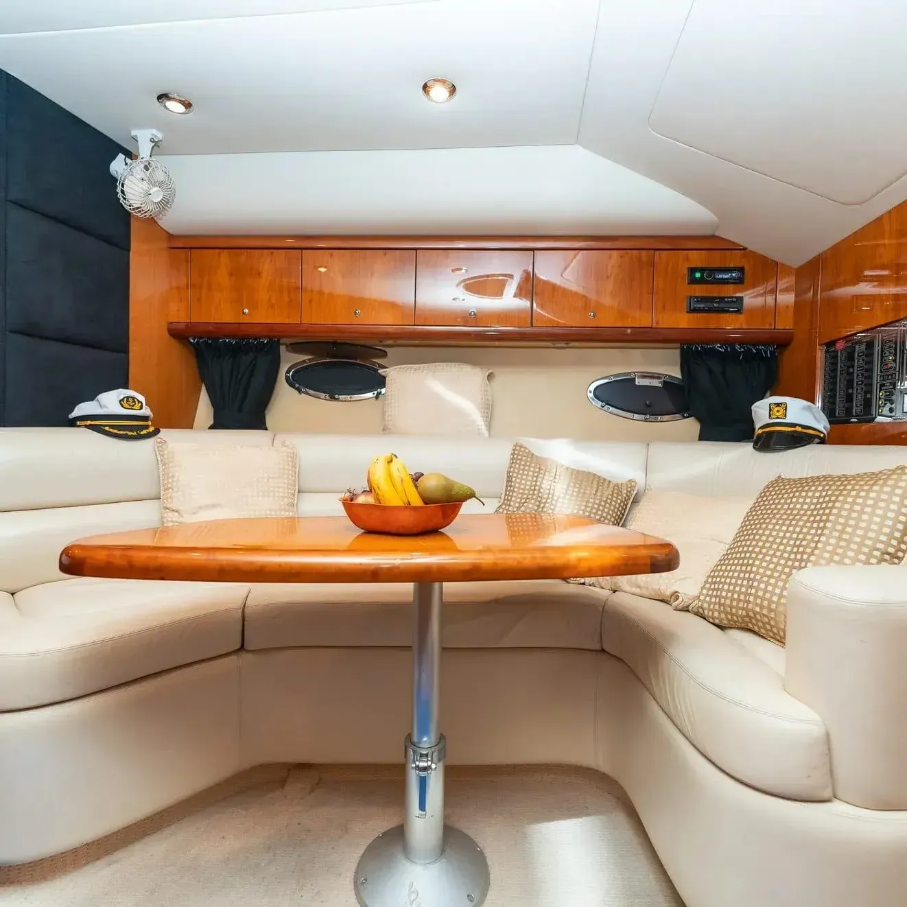 Interior de un yate con sofá modular blanco roto, mesa de madera y frutero. Sombreros de capitán a ambos lados.