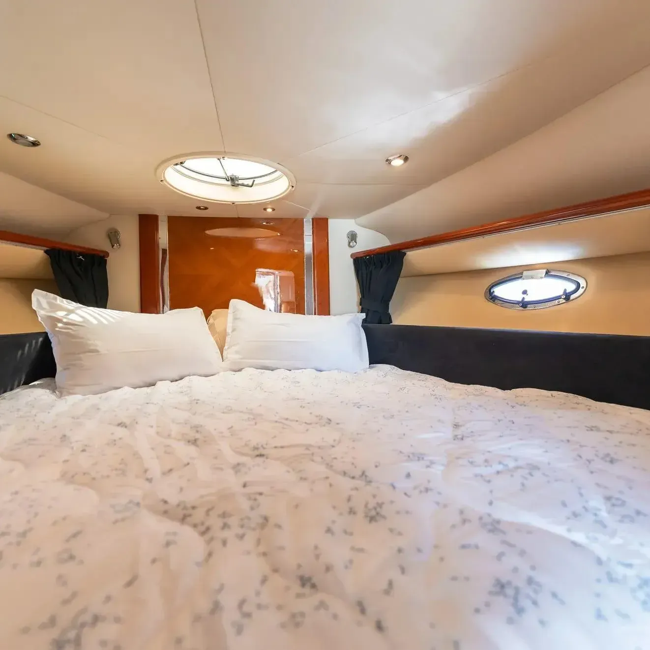 Dormitorio en un barco con una cama cubierta con un edredón y almohadas blancas. Hay luces en el techo y un ojo de buey.