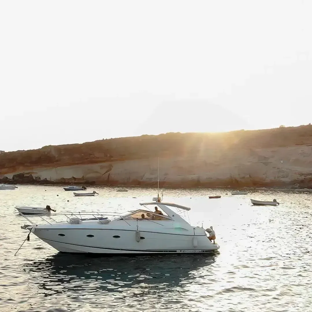 Lancha motora blanca en aguas tranquilas cerca de una costa rocosa, con el sol poniéndose al fondo.