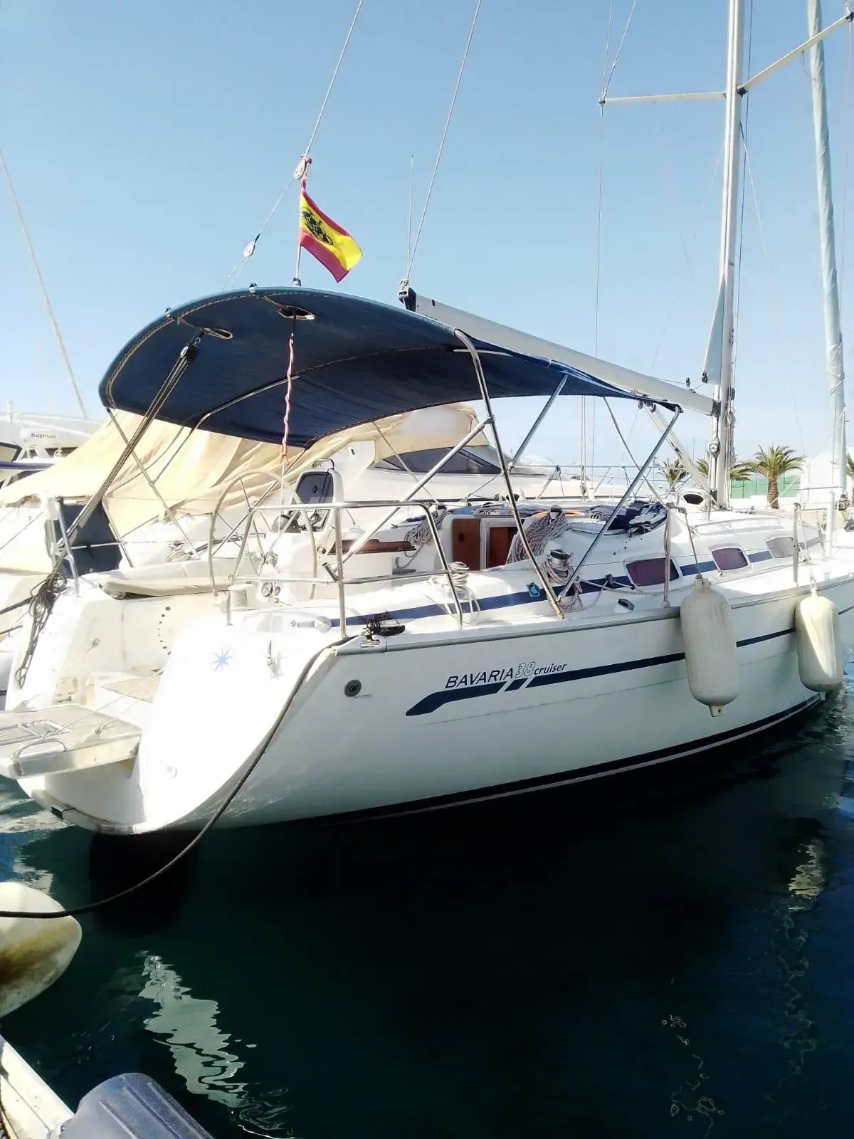 Velero con capota azul y bandera española, atracado en un puerto deportivo en un día soleado.