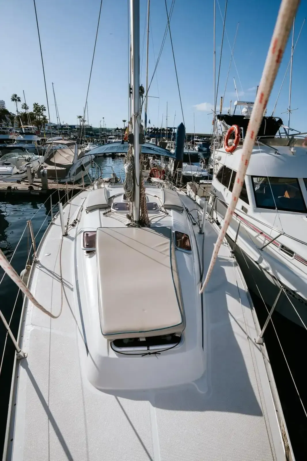 Un velero blanco atracado en un puerto deportivo, visto desde arriba, con mástiles, aparejos y un cielo azul sobre sus cabezas.