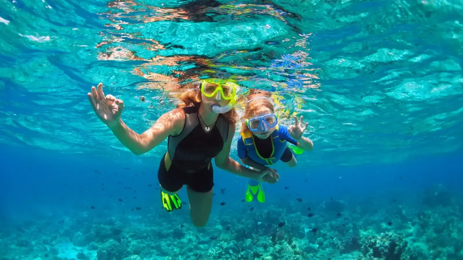 Mujer y niño haciendo snorkel en aguas cristalinas, haciendo señales de OK, sobre un arrecife de coral.