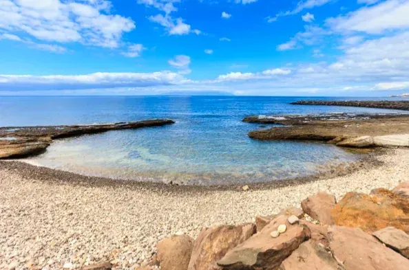 Escena de playa con agua azul tranquila, costa de guijarros y cielo azul brillante con nubes.