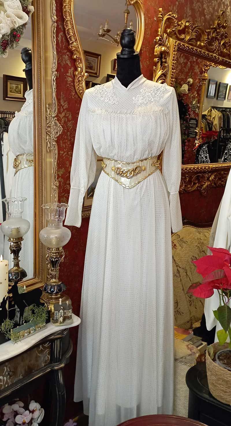 Vestido vintage blanco sobre maniquí con cinturón dorado, expuesto en una tienda con decoración antigua y espejo.