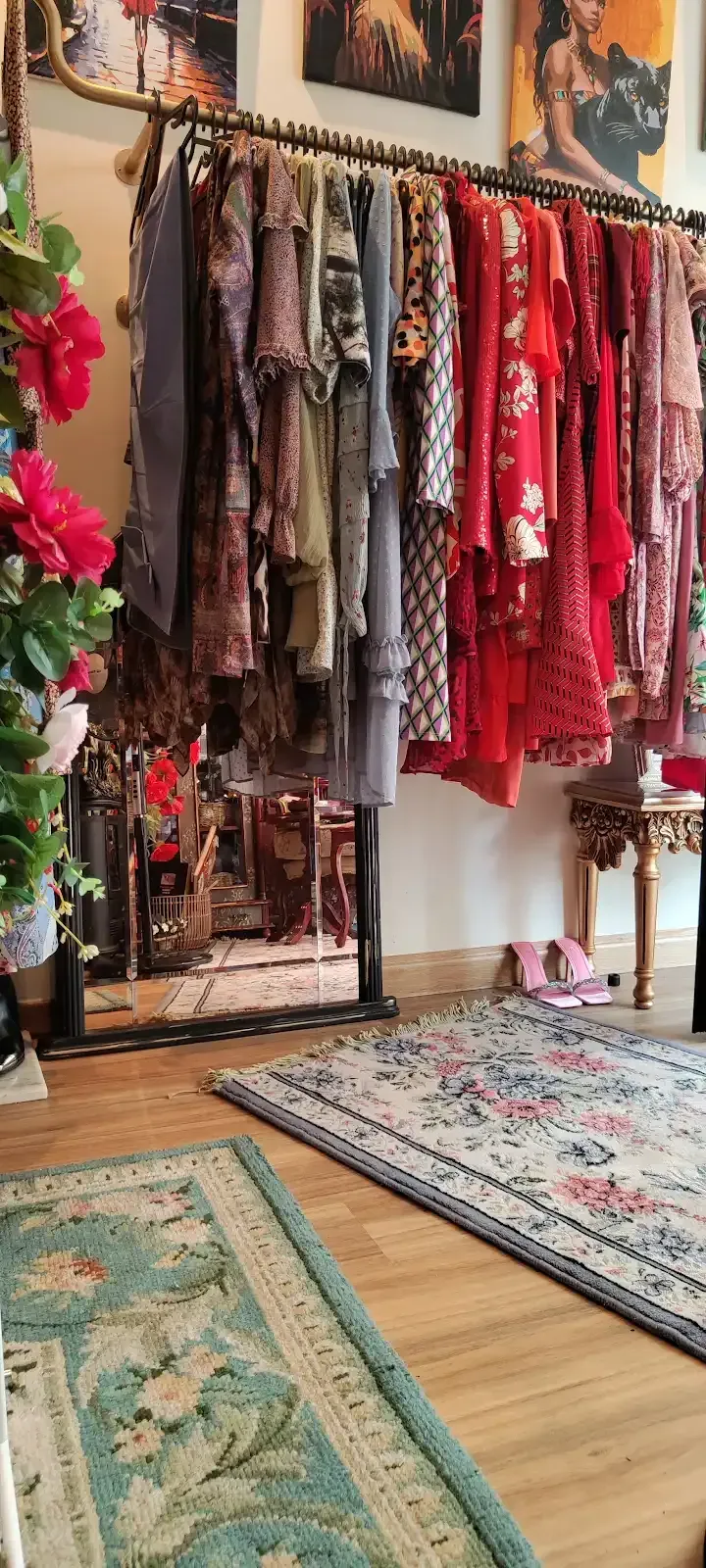 Perchero con vestidos en una boutique. Dos alfombras sobre suelo de madera. Flores rosas en la esquina izquierda.