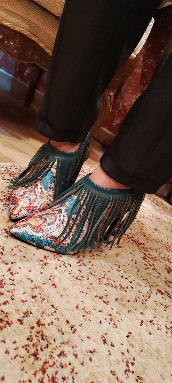 Persona que viste pantalones negros y zapatos con flecos de color verde azulado sobre una alfombra estampada.