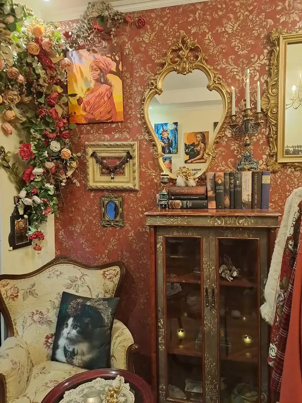 Habitación acogedora con arreglos florales, espejo ornamentado, armario y silla con cojín de gato. Paredes rojas.
