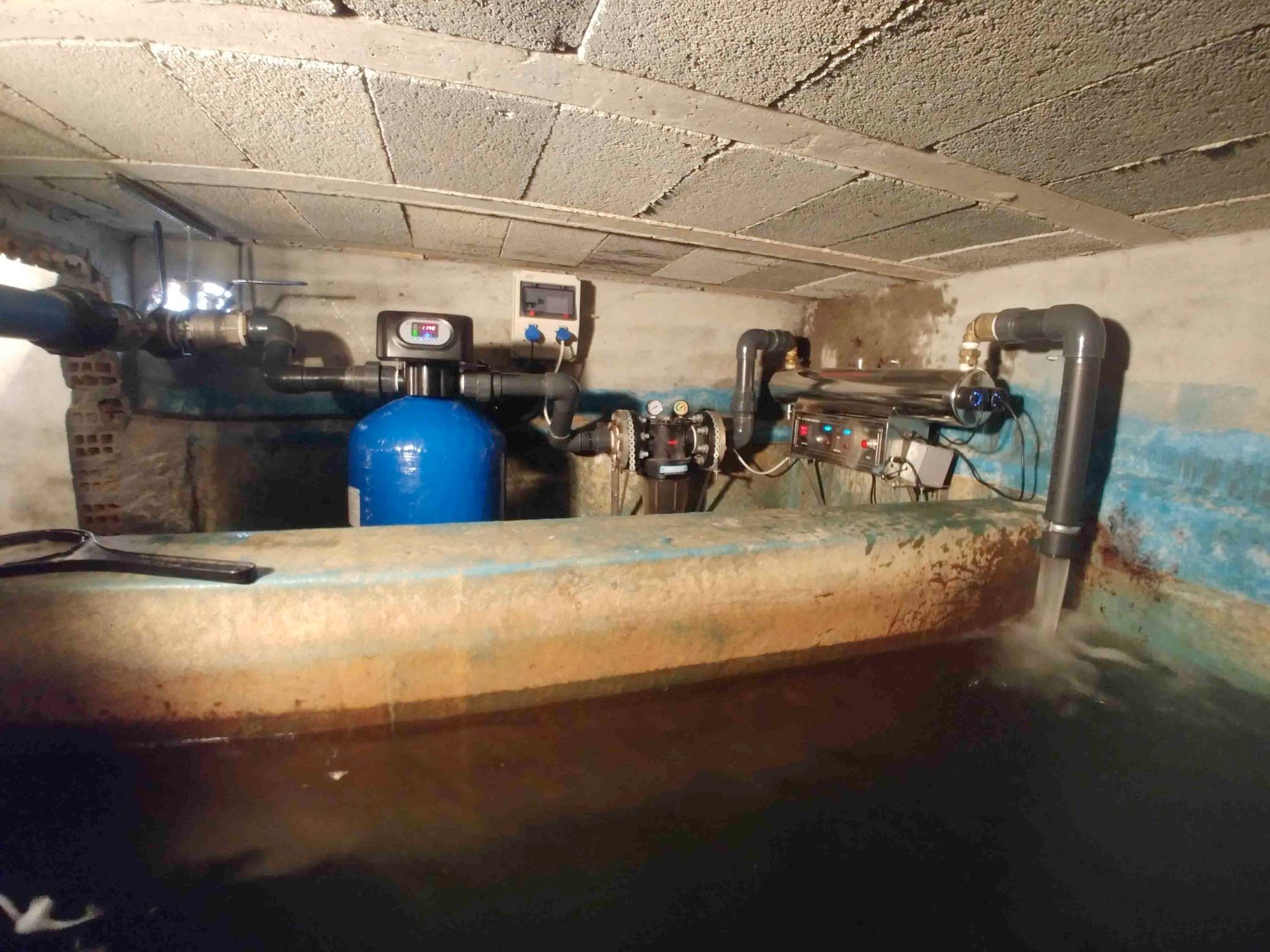 Sala de tratamiento de agua industrial con tanques, tuberías y maquinaria en un sótano estrecho de hormigón