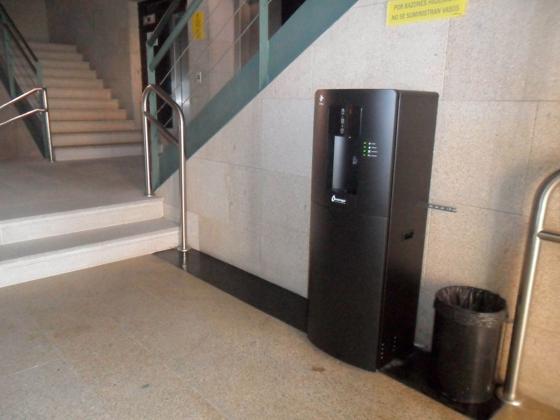 Vestíbulo interior con escaleras, una máquina expendedora negra y una papelera junto a la pared