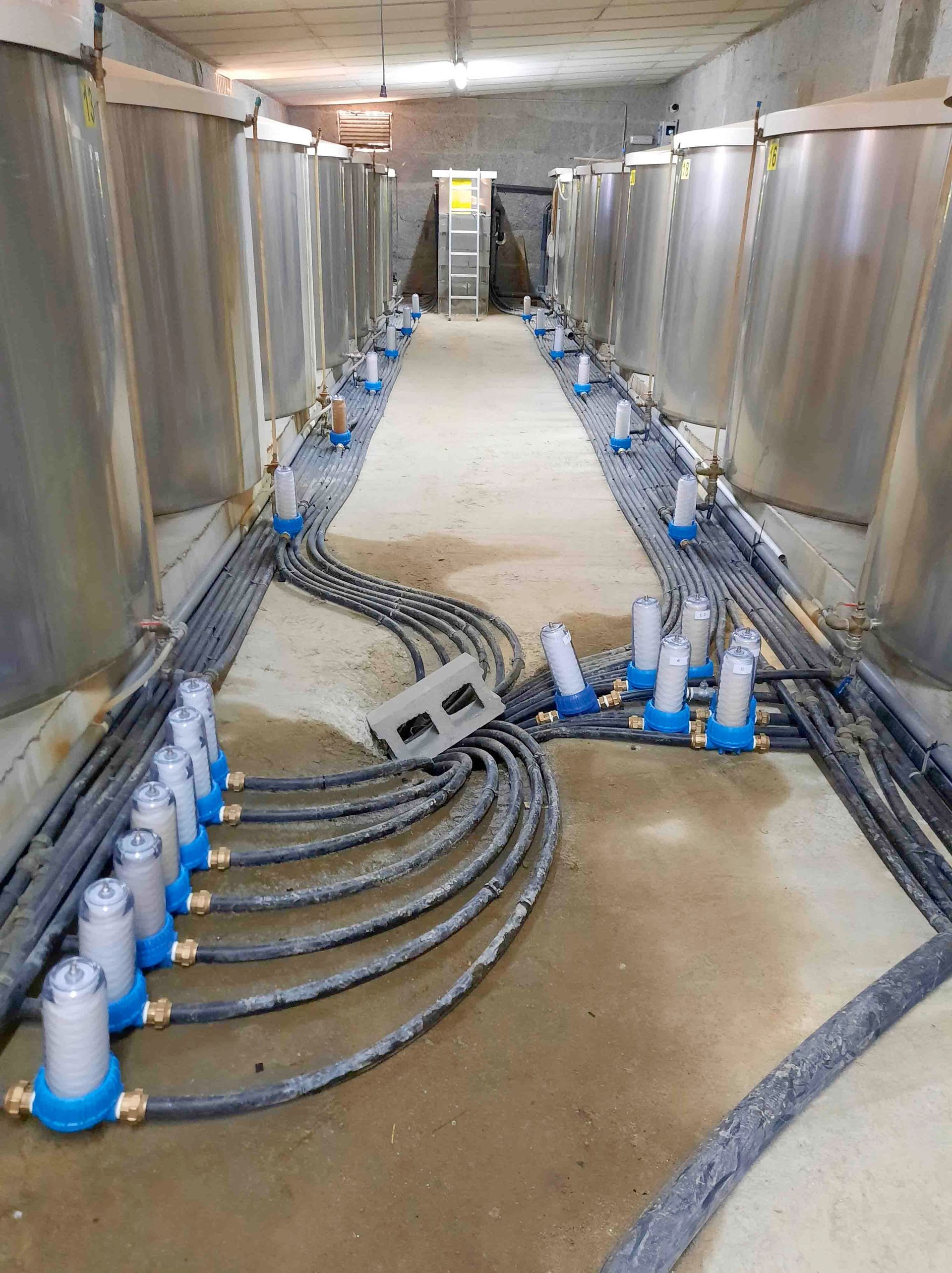 Pasillo industrial con filas de grandes tanques metálicos y cables agrupados en el suelo