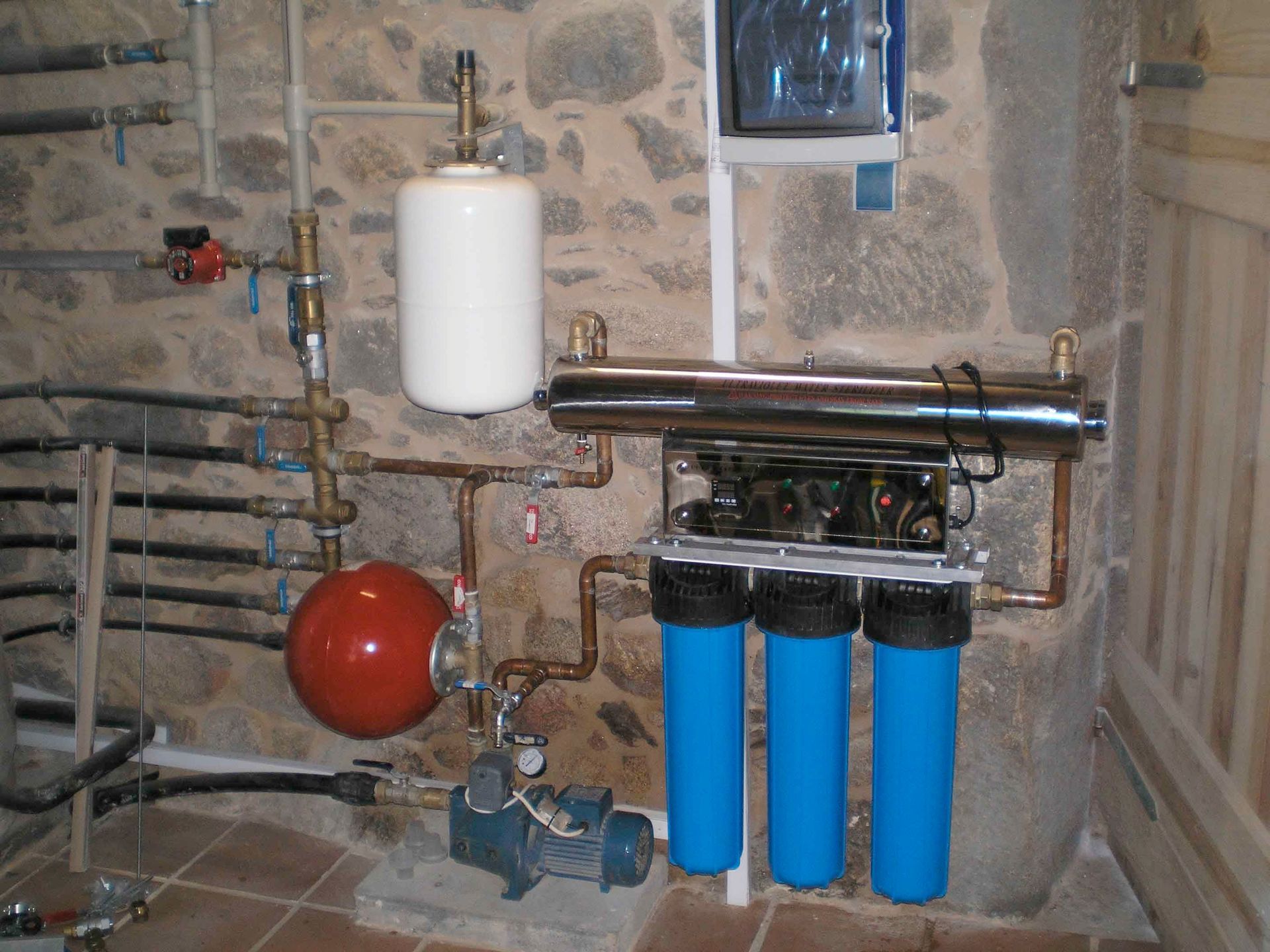 Sistema de filtración de agua en el sótano, con carcasas de filtro azules, tuberías y un tanque de presión rojo, junto a una pared de piedra