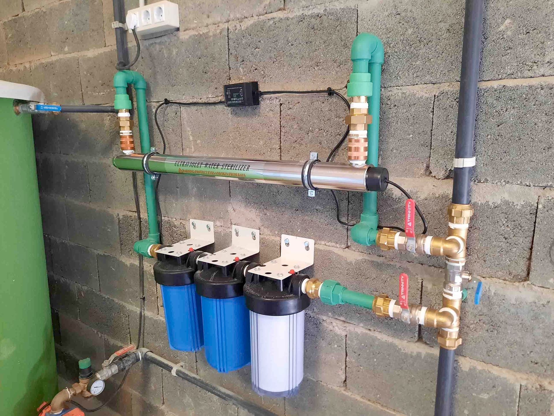 Tres carcasas de filtros de agua y tuberías de PVC instaladas en una pared de bloques de hormigón, cerca de un depósito verde.