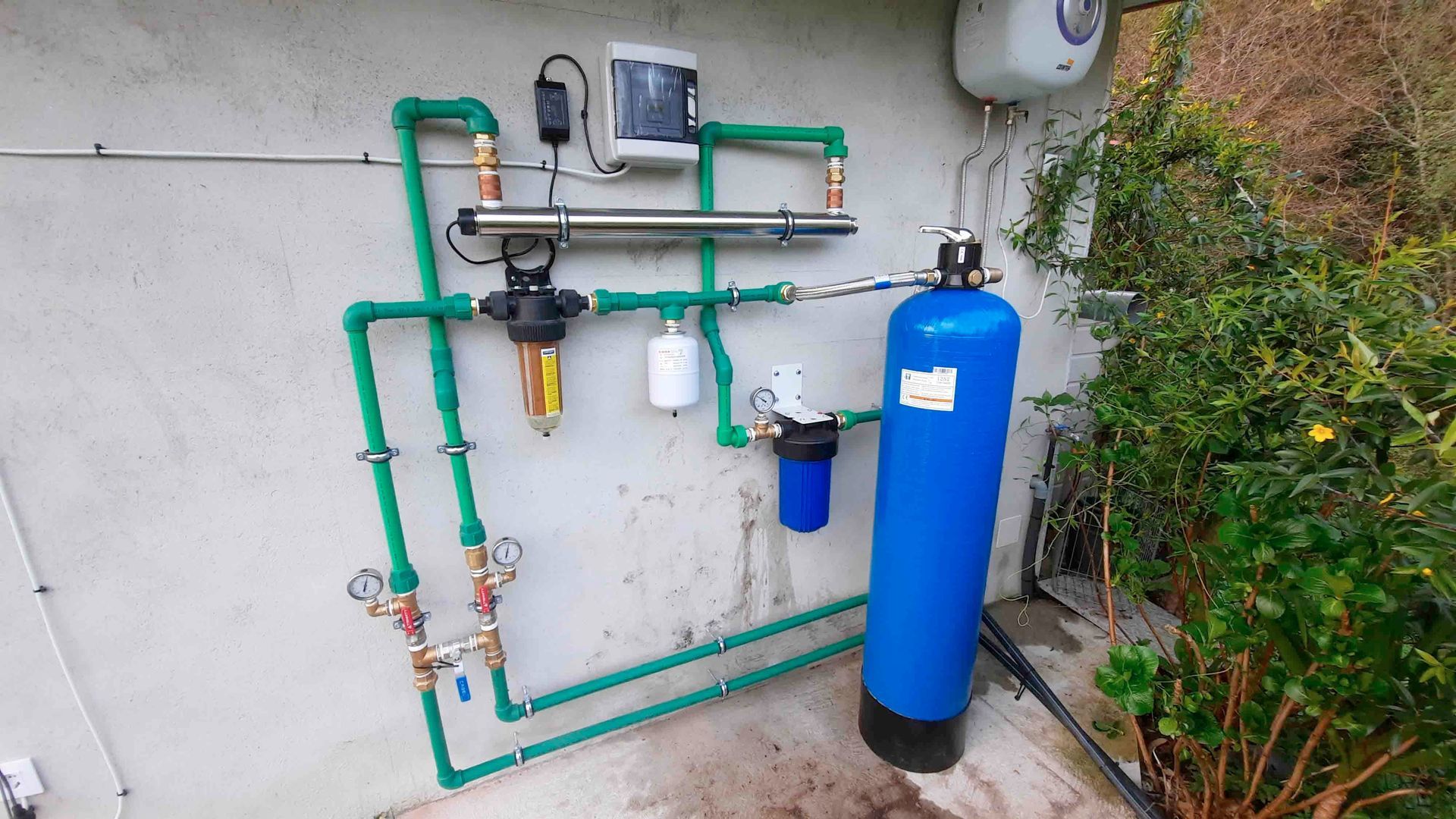 Sistema de filtración de agua azul con tuberías verdes instalado en una pared exterior junto a unos arbustos