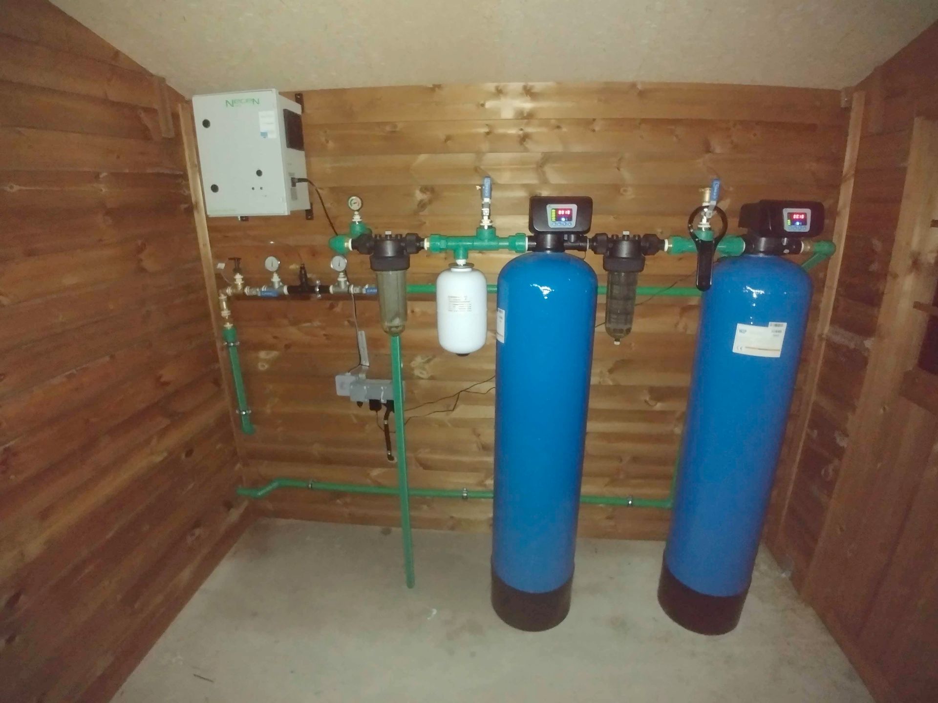 Equipos de tratamiento de agua en un cuarto de servicio de madera, con dos grandes depósitos azules y tuberías.