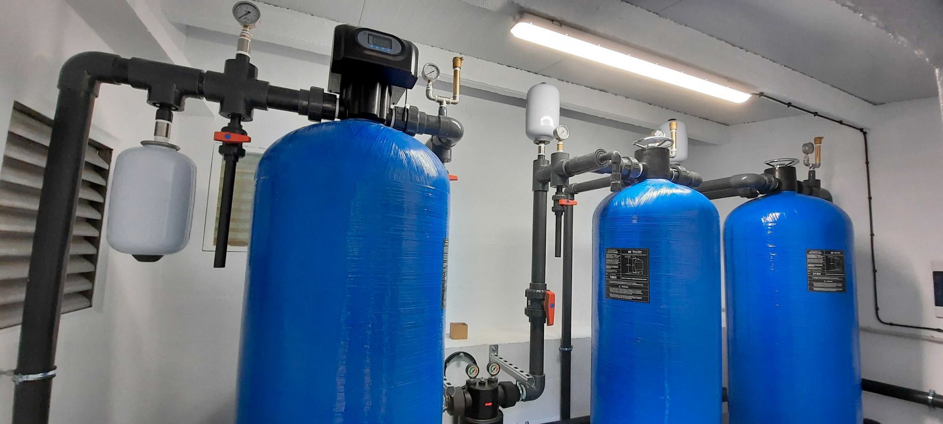 Depósitos y tuberías azules de filtración de agua en un cuarto de servicio con techo blanco e iluminación