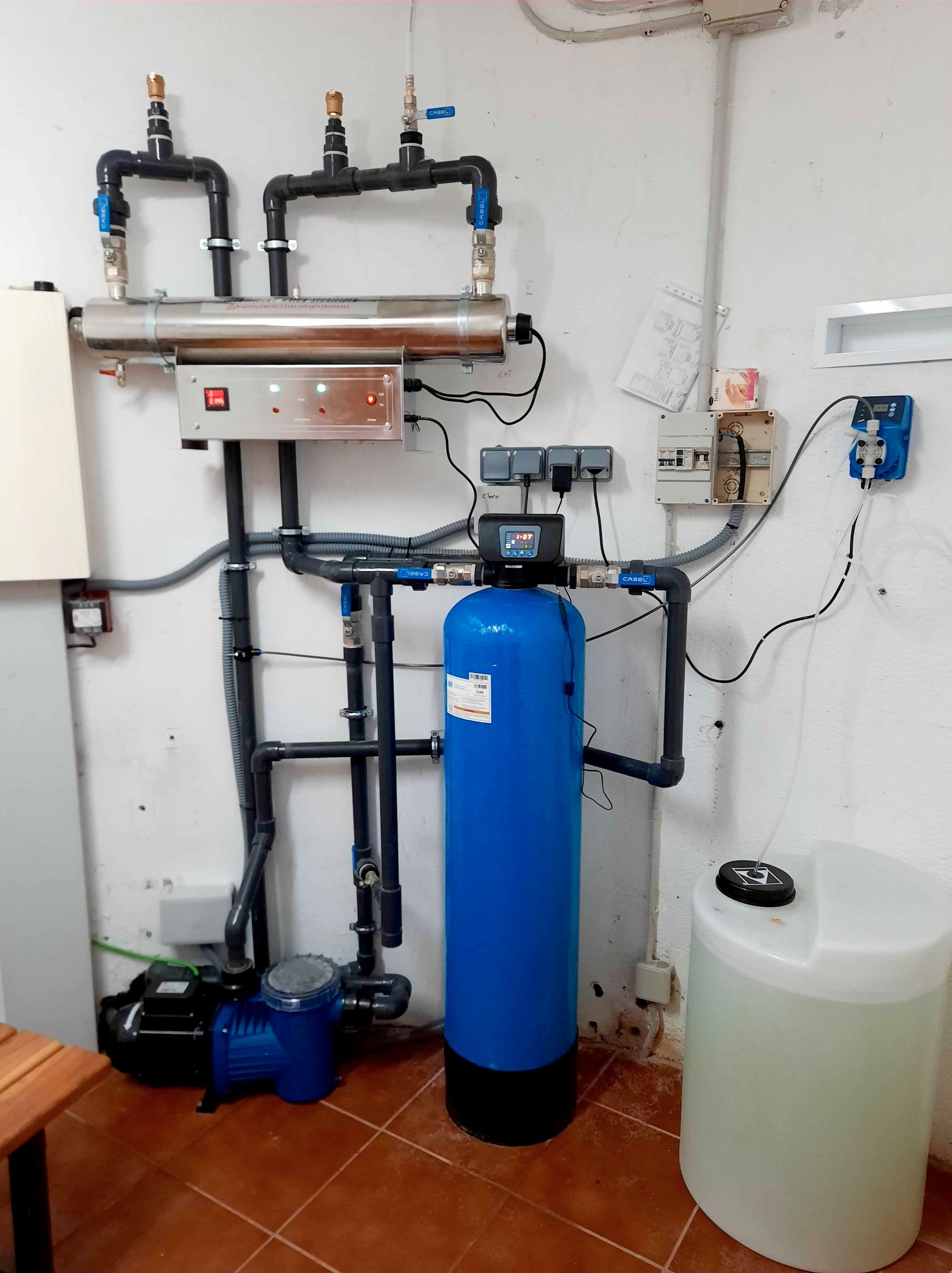 Equipo de tratamiento de agua: tanque de filtración azul, tuberías, bomba y tanque de almacenamiento blanco en un cuarto de servicio
