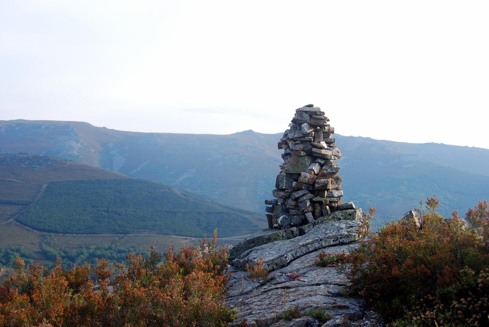 Mojón de piedras en un mirador de montaña con arbustos otoñales y crestas azules en la distancia.