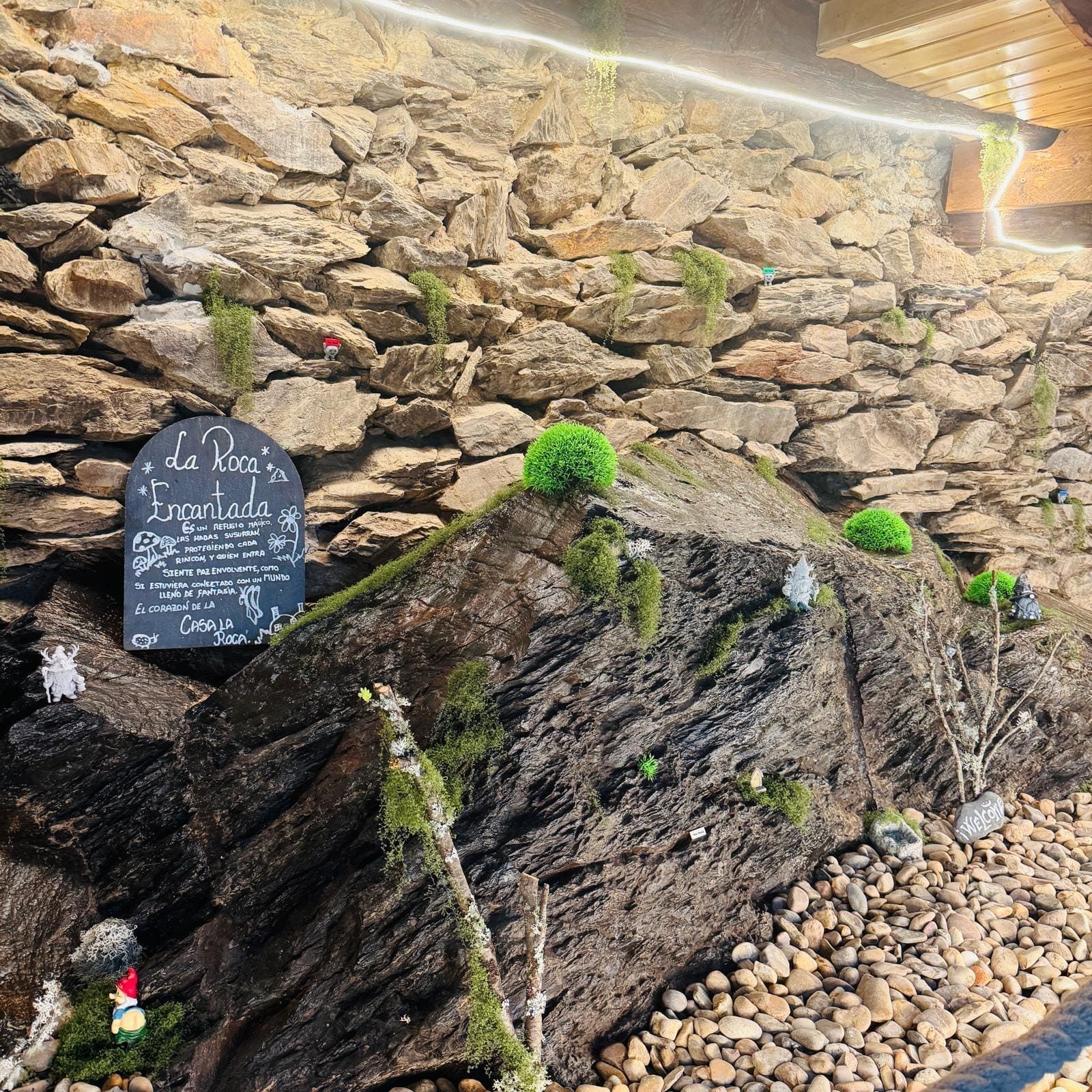 Muro rocoso de jardín con musgo y un pequeño cartel de pizarra sobre un fondo de piedra.
