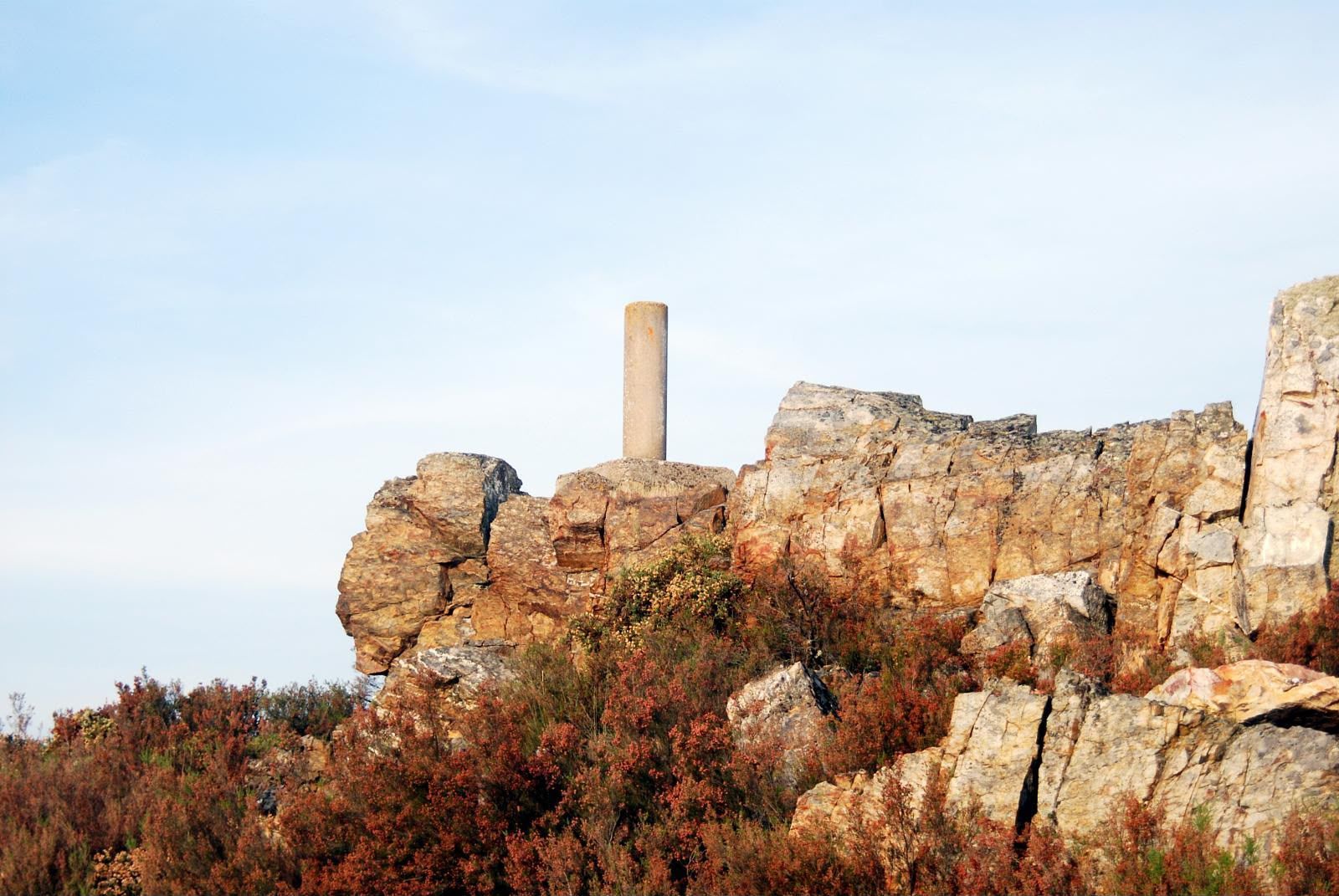 Acantilado rocoso con antiguas ruinas de piedra y un pilar solitario sobre arbustos otoñales bajo un cielo azul pálido.