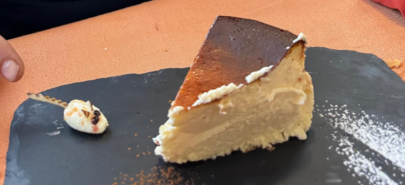 Una porción de tarta de queso en un plato de pizarra negra con una cucharada de crema sobre una mesa naranja.