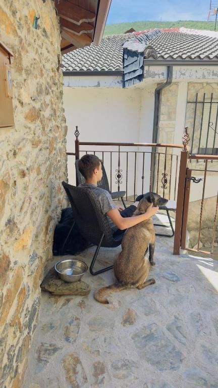 Una persona se relaja en una silla en un patio de piedra junto a un perro pequeño, con tejados al fondo.