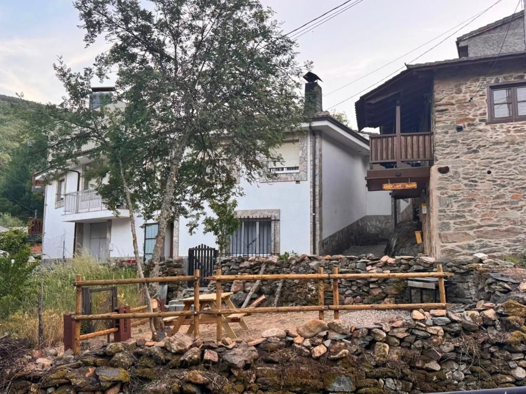 Casa de piedra y edificio blanco con un árbol delante, junto a una valla de madera y un muro de piedra en un pueblo rural.