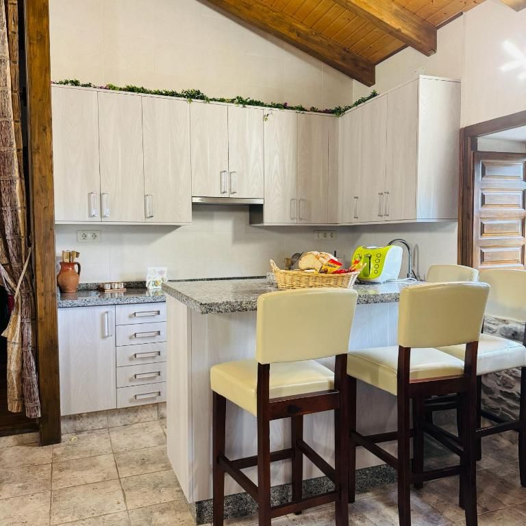 Cocina luminosa con armarios blancos, isla de granito, dos taburetes de bar color crema y techo con vigas de madera.