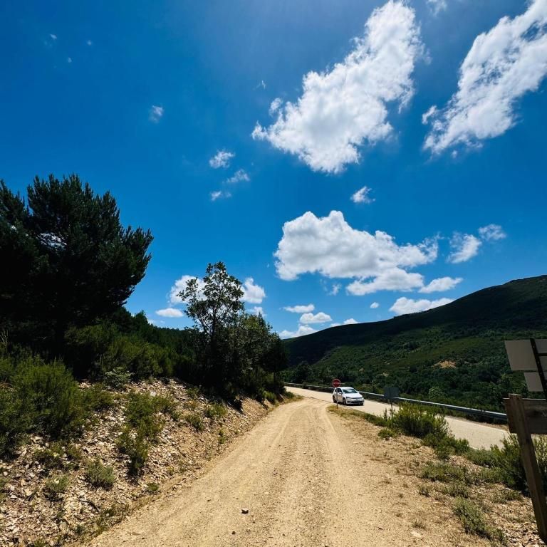 Camino de tierra que atraviesa verdes colinas bajo un cielo azul brillante con nubes, con un coche blanco aparcado más adelante.