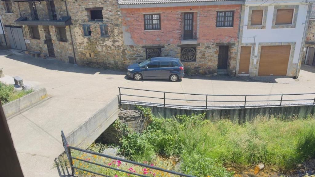 Calle tranquila con un coche azul aparcado junto a antiguos edificios de ladrillo y una barandilla sobre vegetación exuberante.