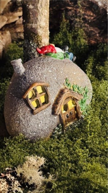 Casita de piedra en miniatura con ventanas de un amarillo brillante y techo rojo, enclavada entre musgo y vegetación.