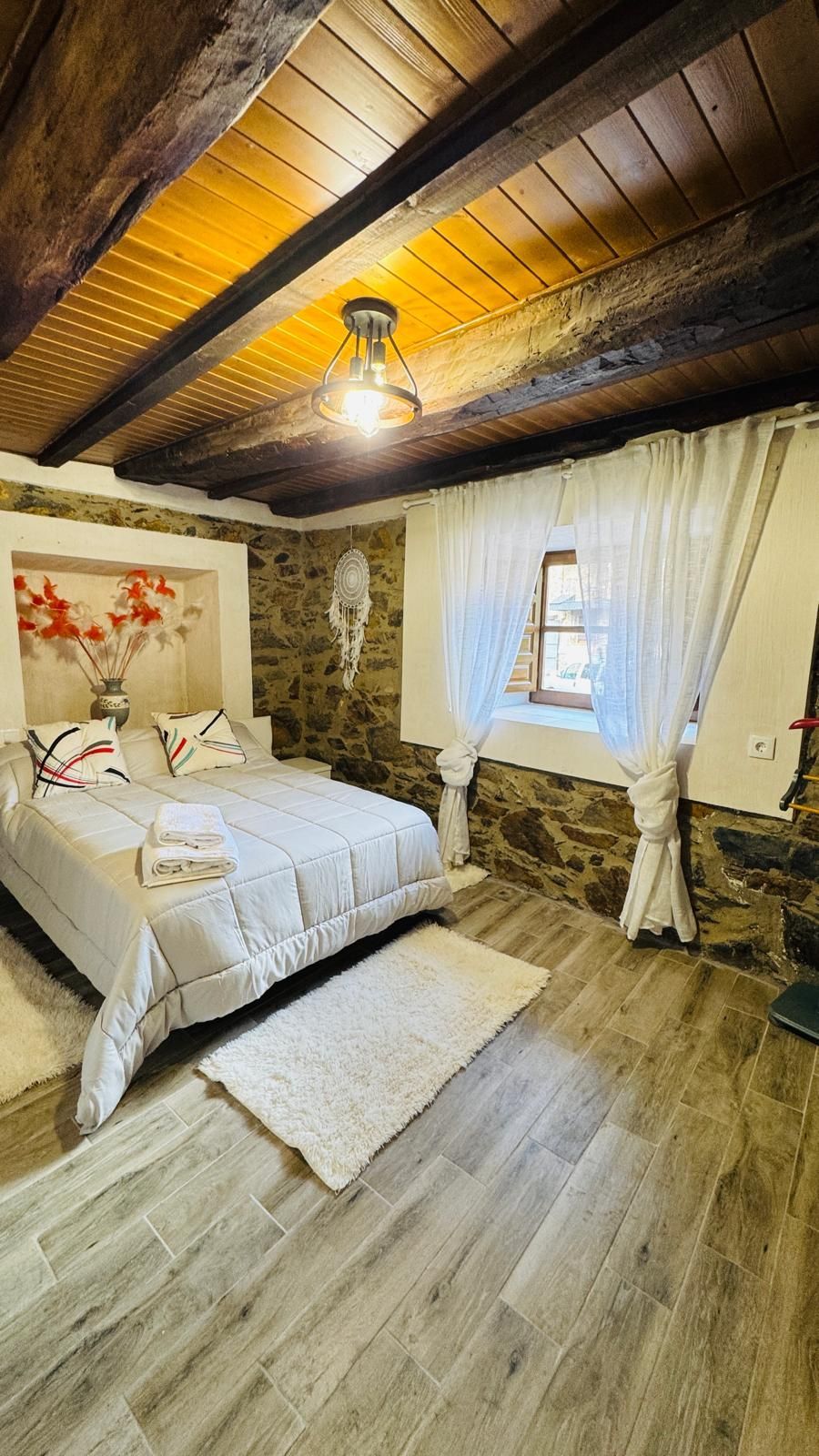 Acogedora habitación rústica con techo de madera, cama blanca, ventana con cortinas e iluminación cálida.