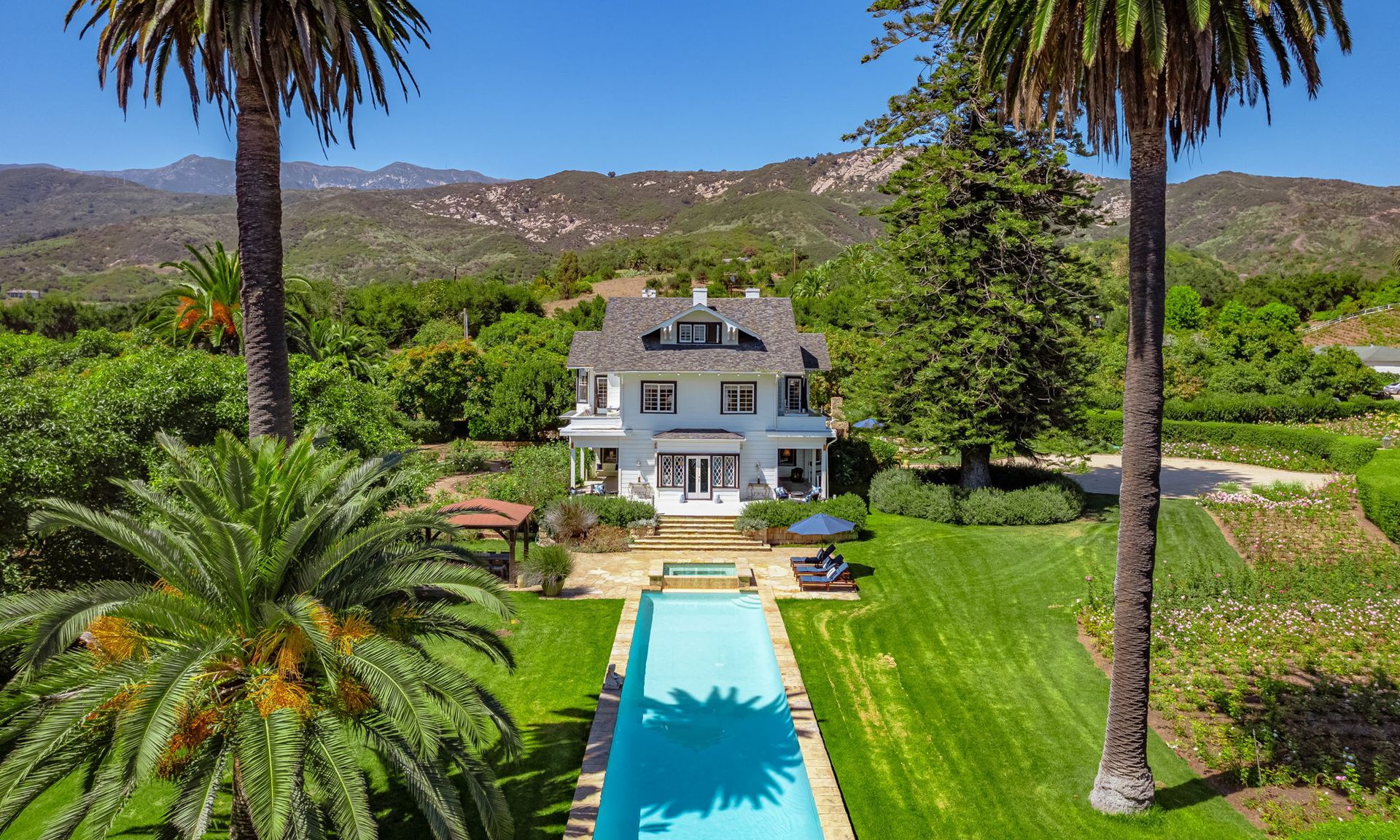 Modern white house with a long turquoise pool, lush lawn, palm trees, and mountain backdrop