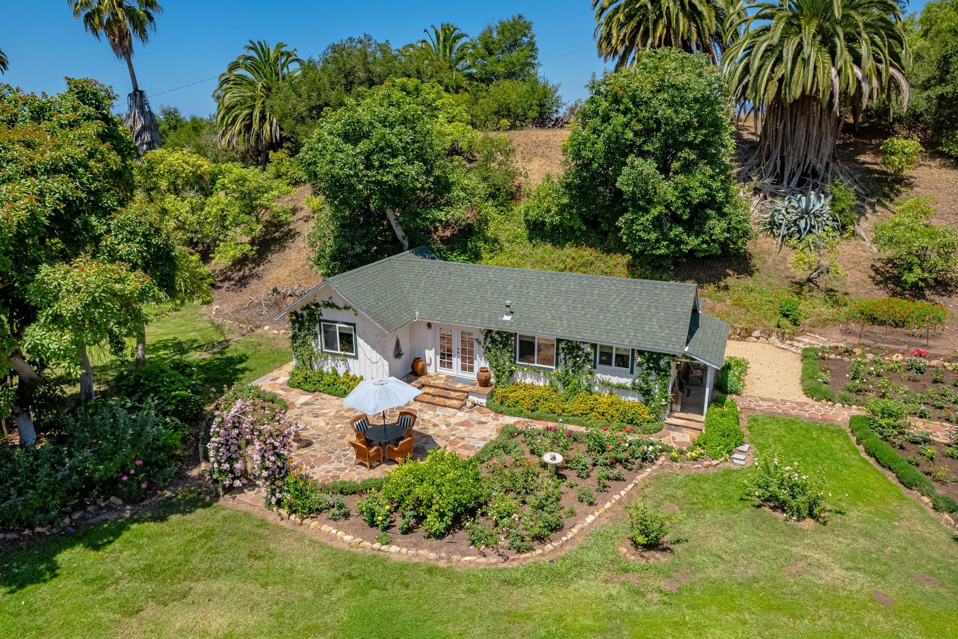 Aerial view of a small house with a patio, surrounded by lush green trees and hillside landscaping.