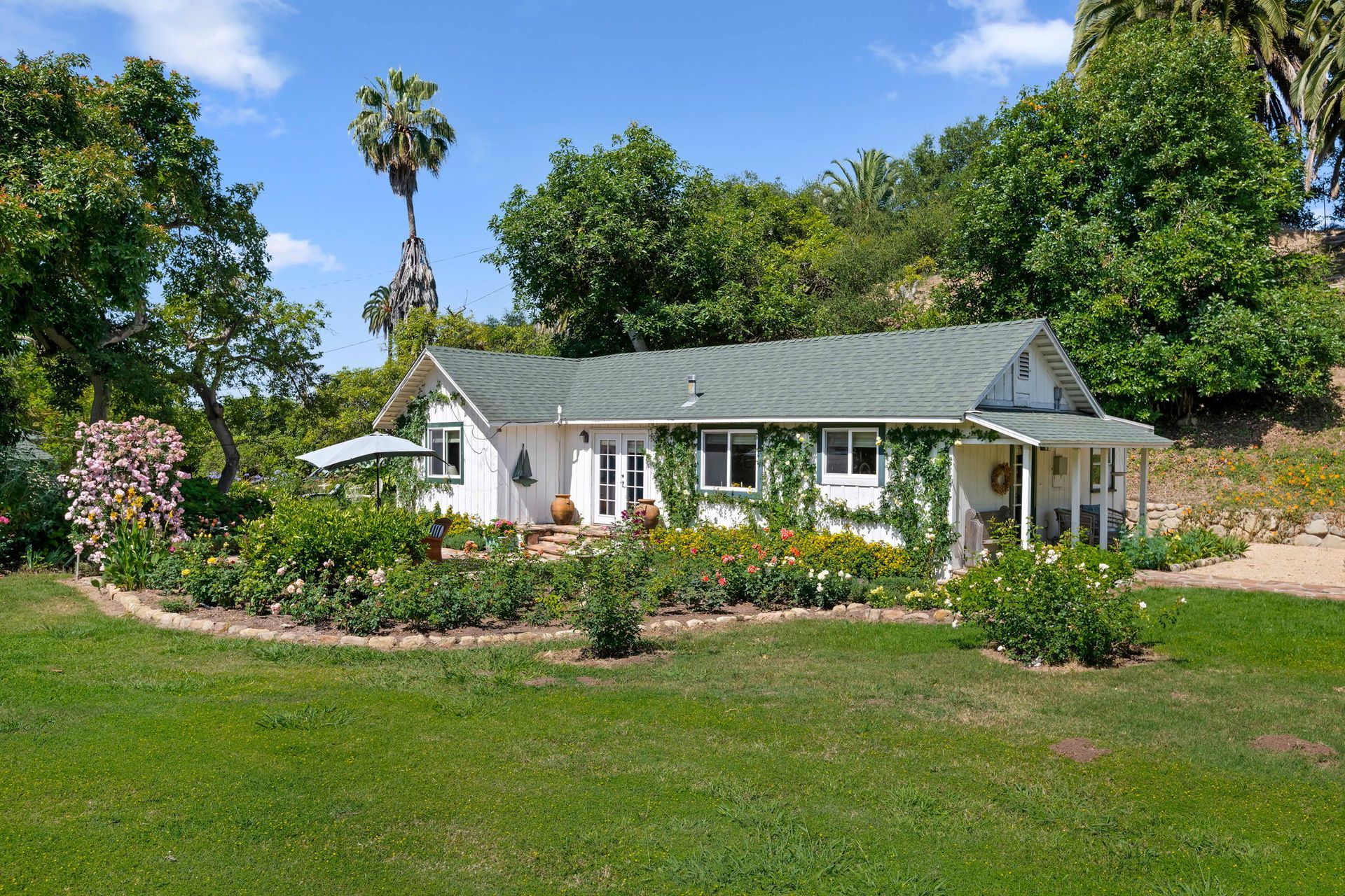 White cottage with a green roof and flower garden under a blue sky, surrounded by trees and lawn