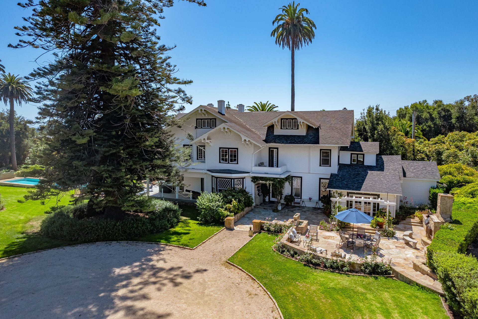 Large white house with a front yard, patio seating, and tall palm trees under a clear blue sky