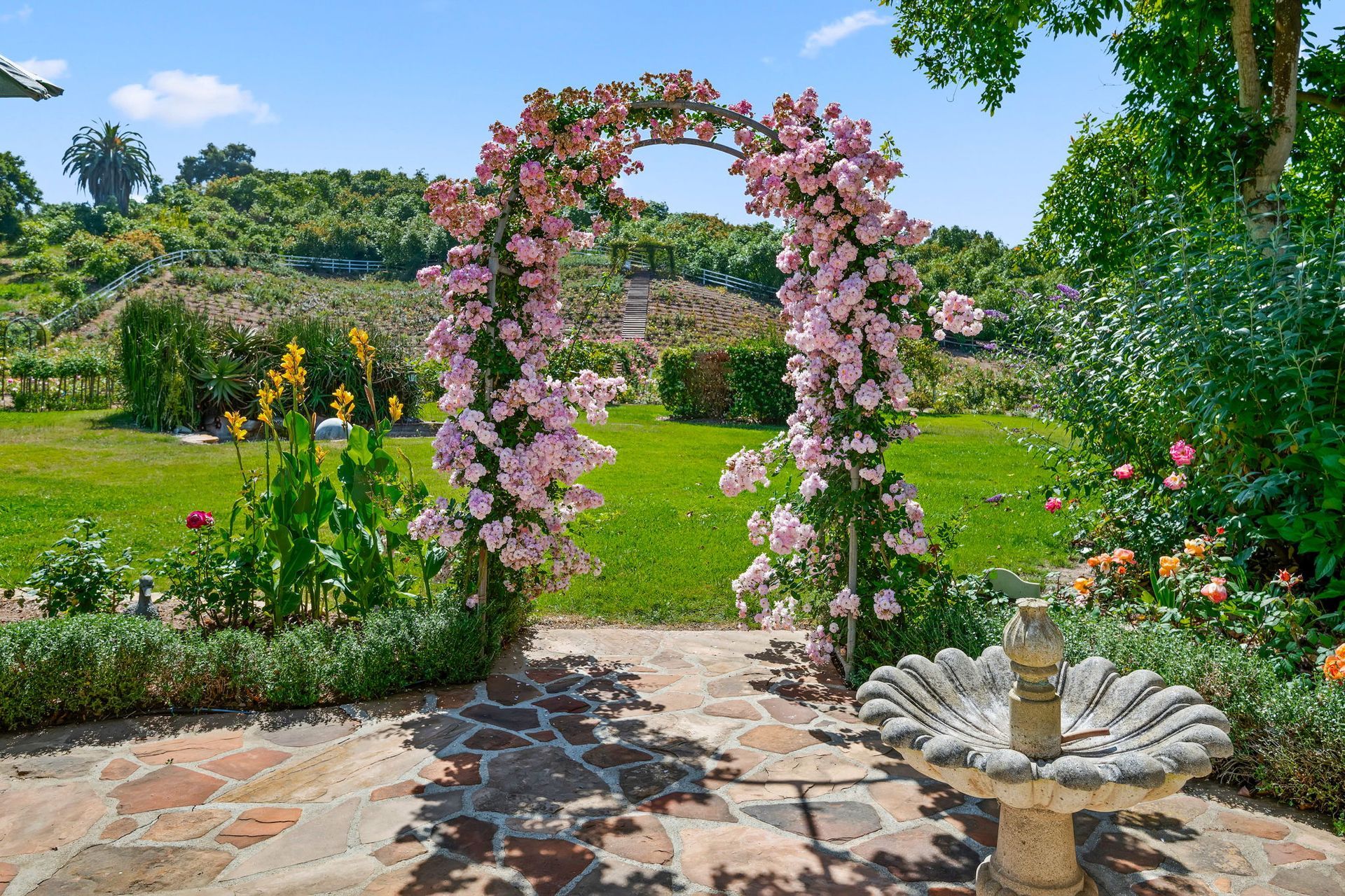 Garden path framed by a pink-flower arch, with a stone fountain and lush green hills in the background