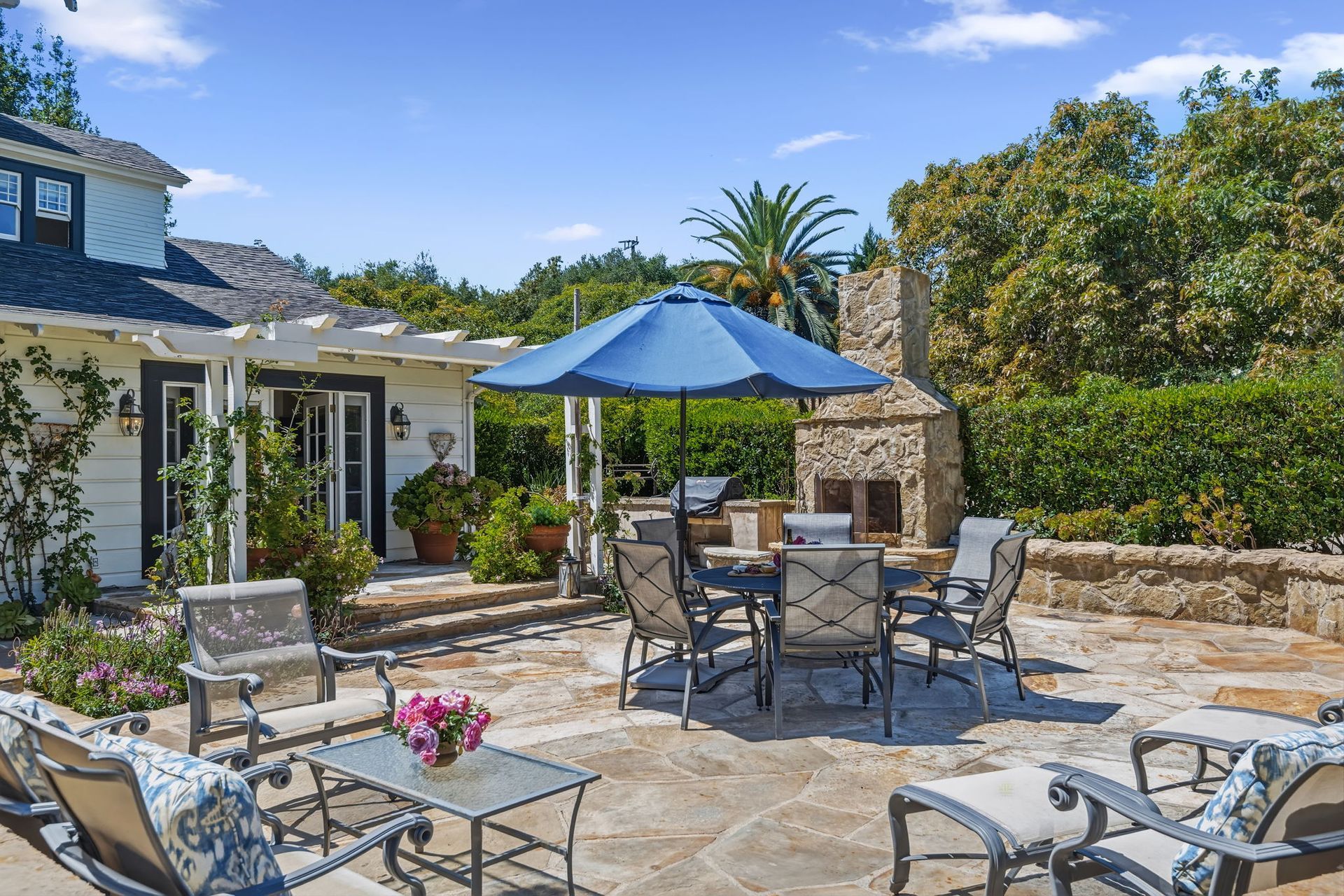 Patio with stone seating, blue umbrella, and outdoor fireplace in a sunny garden courtyard