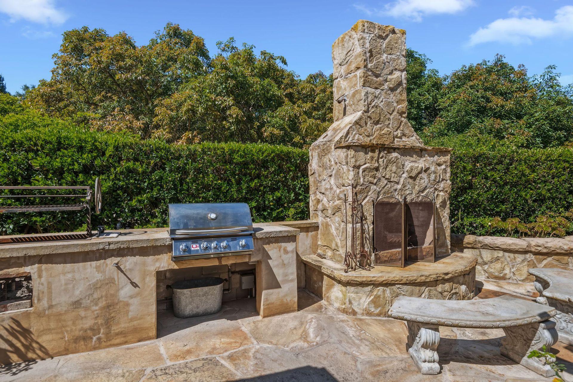 Stone outdoor fireplace with grill and benches on a sunny patio, backed by green trees