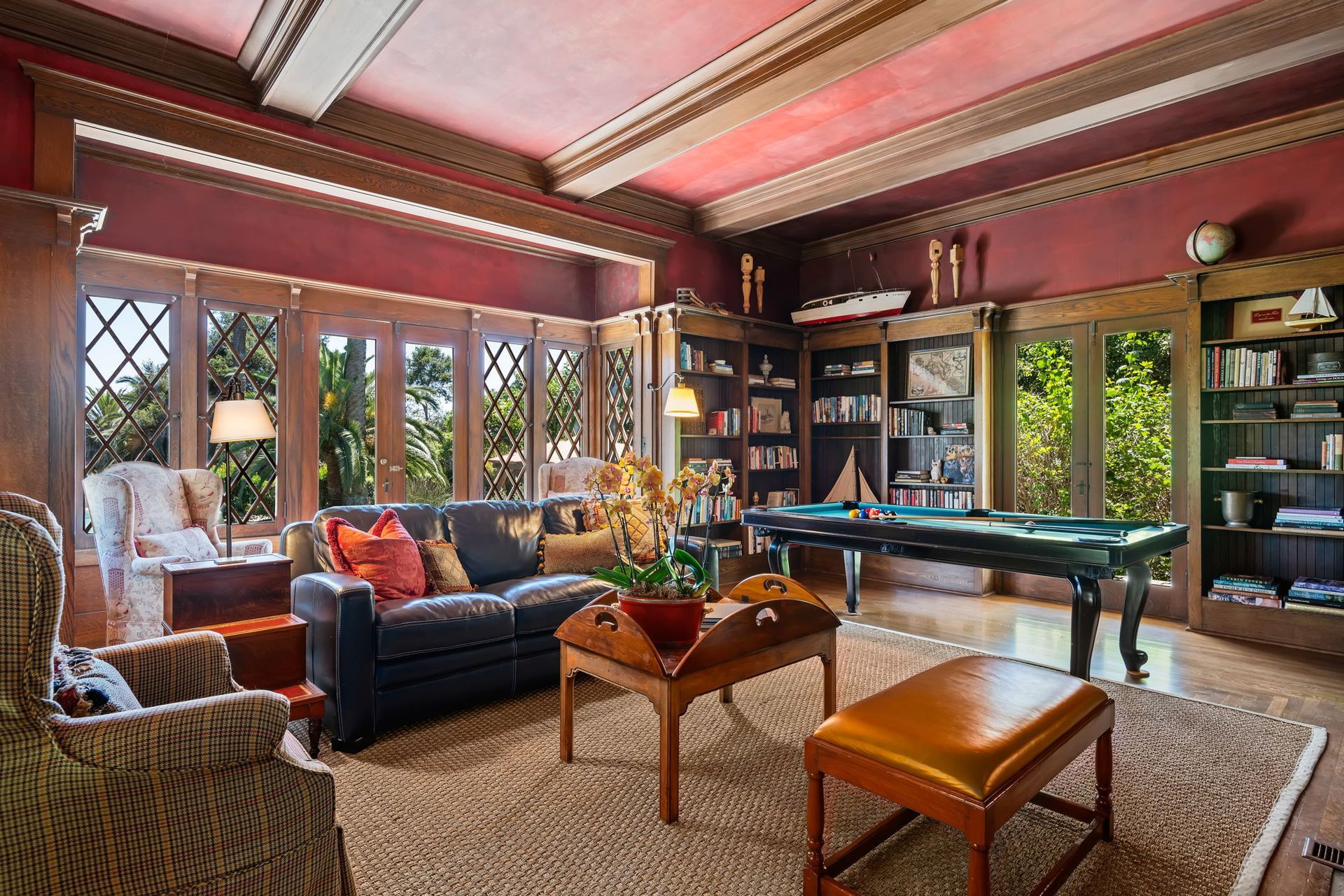 Elegant living room with burgundy walls, black sofa, large windows, bookshelves, and indoor-outdoor garden view