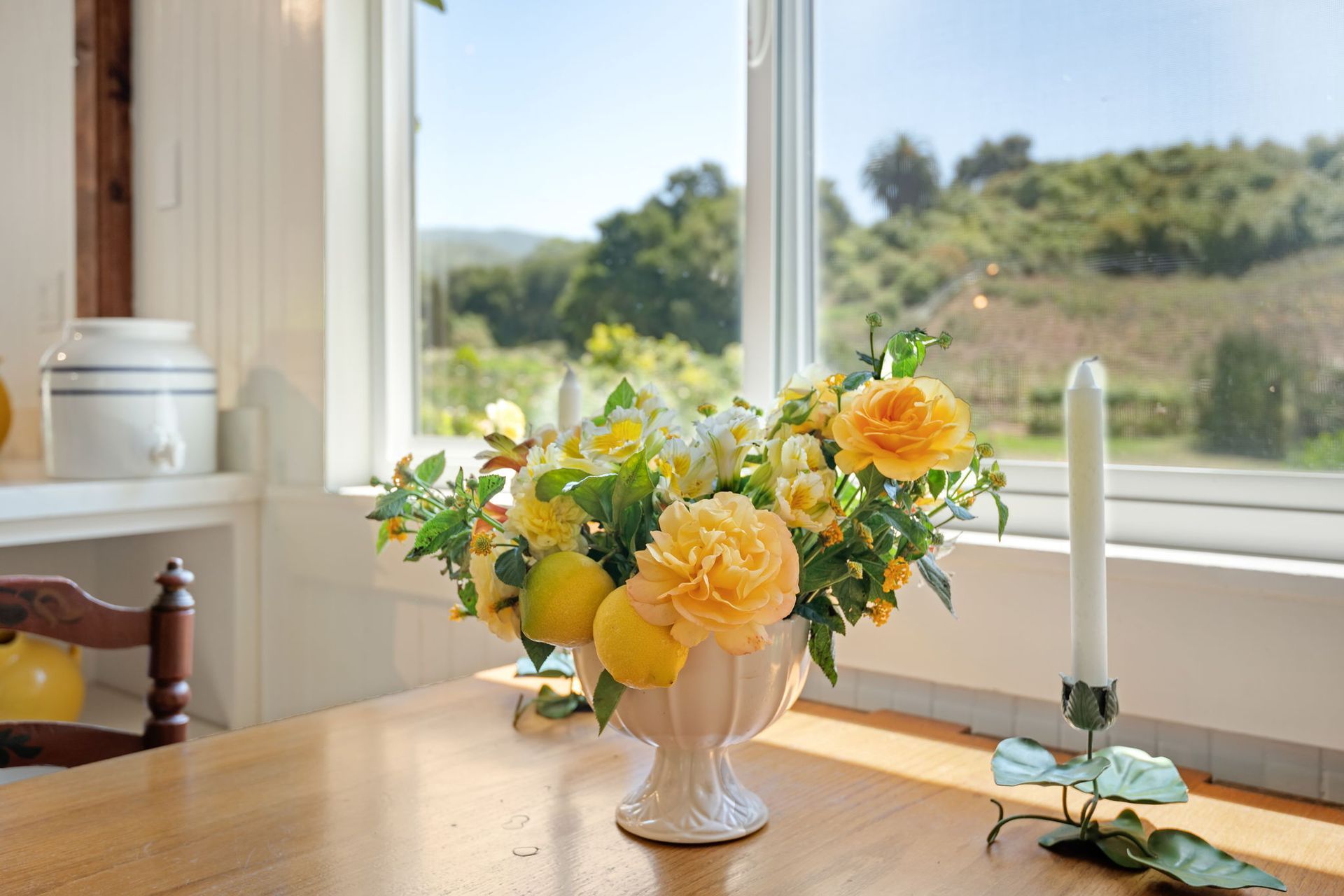 Yellow flowers in a vase on a sunlit wooden table by a window, with a white candle and green hills outside.