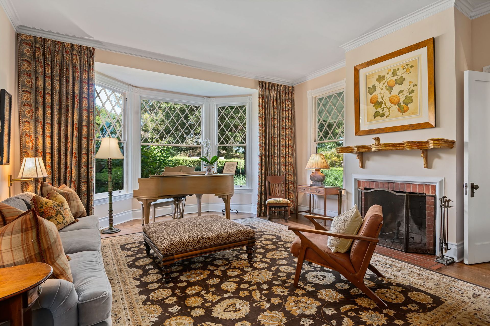 Elegant living room with fireplace, piano, floral decor, patterned rug, and large windows overlooking greenery