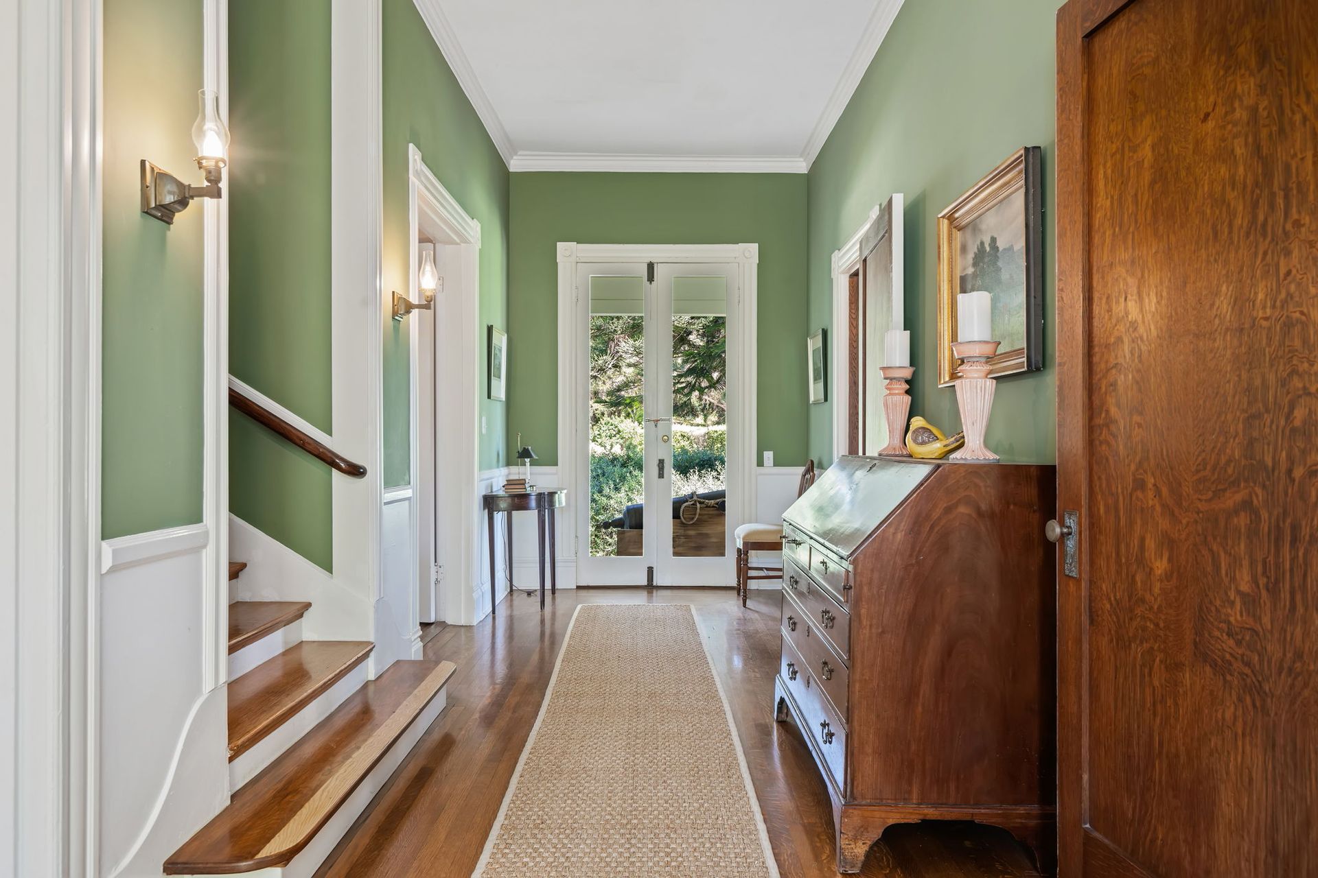 Narrow green hallway with stairs, a runner rug, wooden cabinet, and French doors at the end