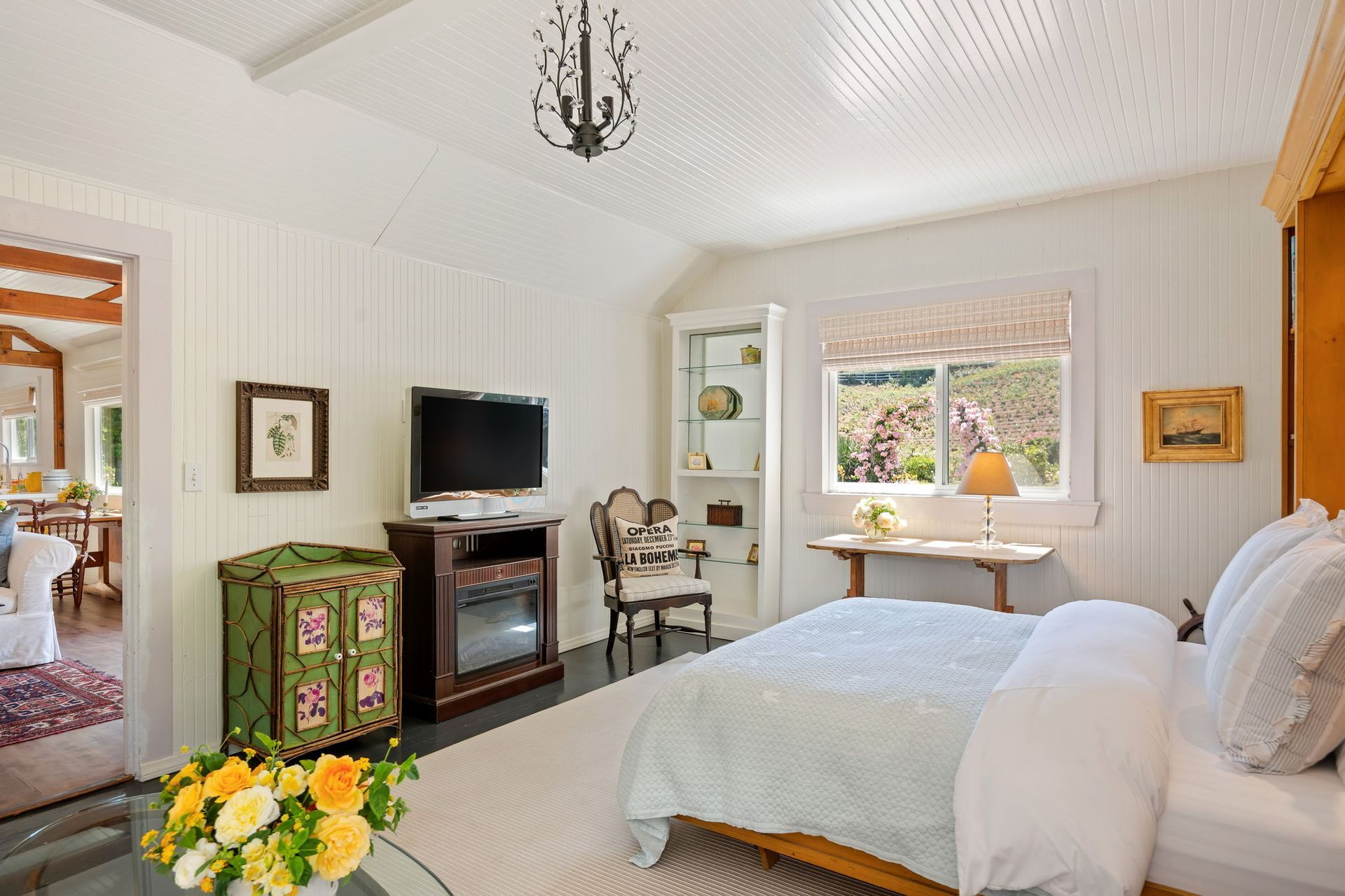 Bright white bedroom with bed, TV, green cabinet, desk by window, and flowers on the floor