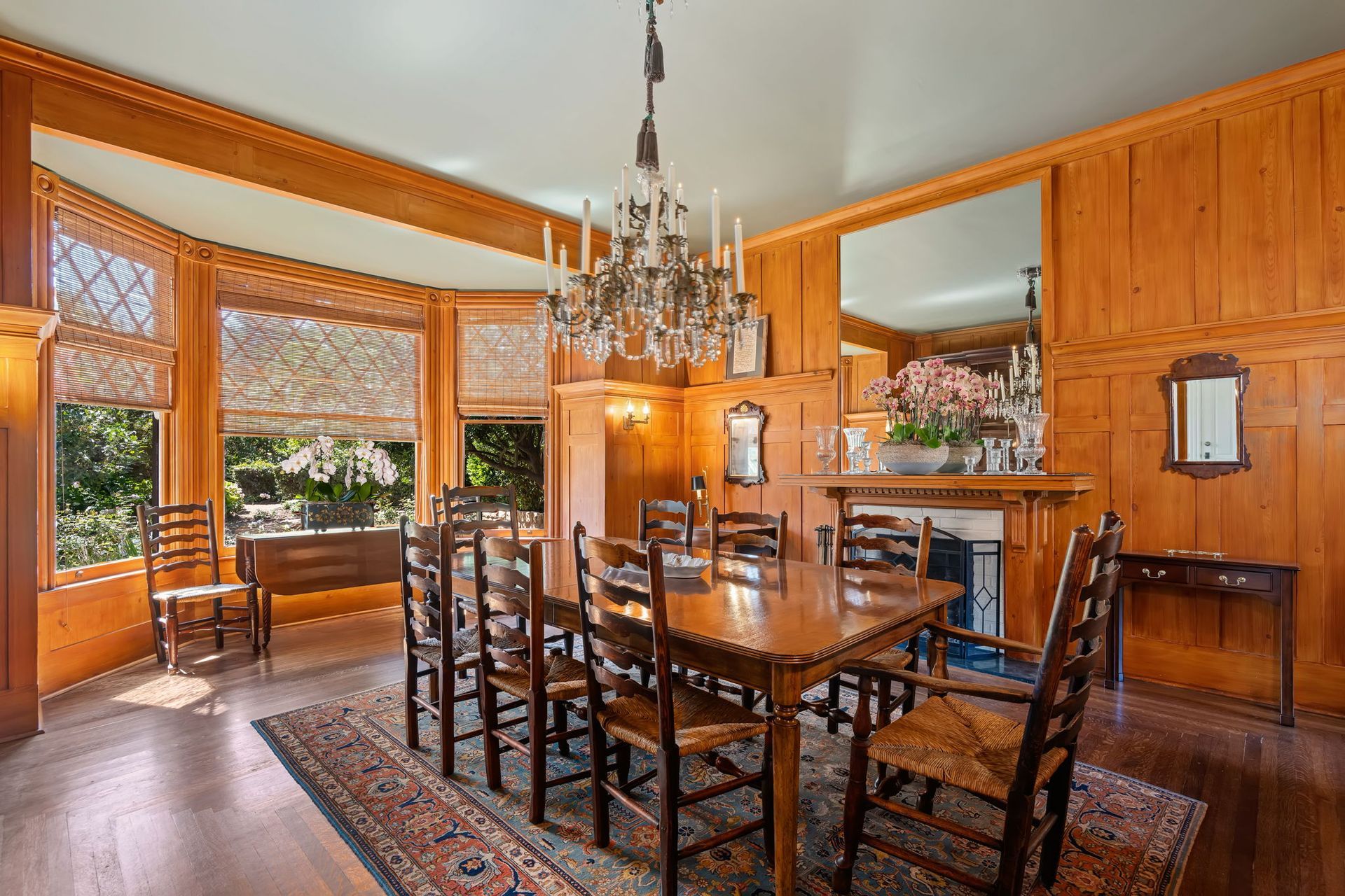 Wood-paneled dining room with chandelier, large table, chairs, and bay windows overlooking greenery