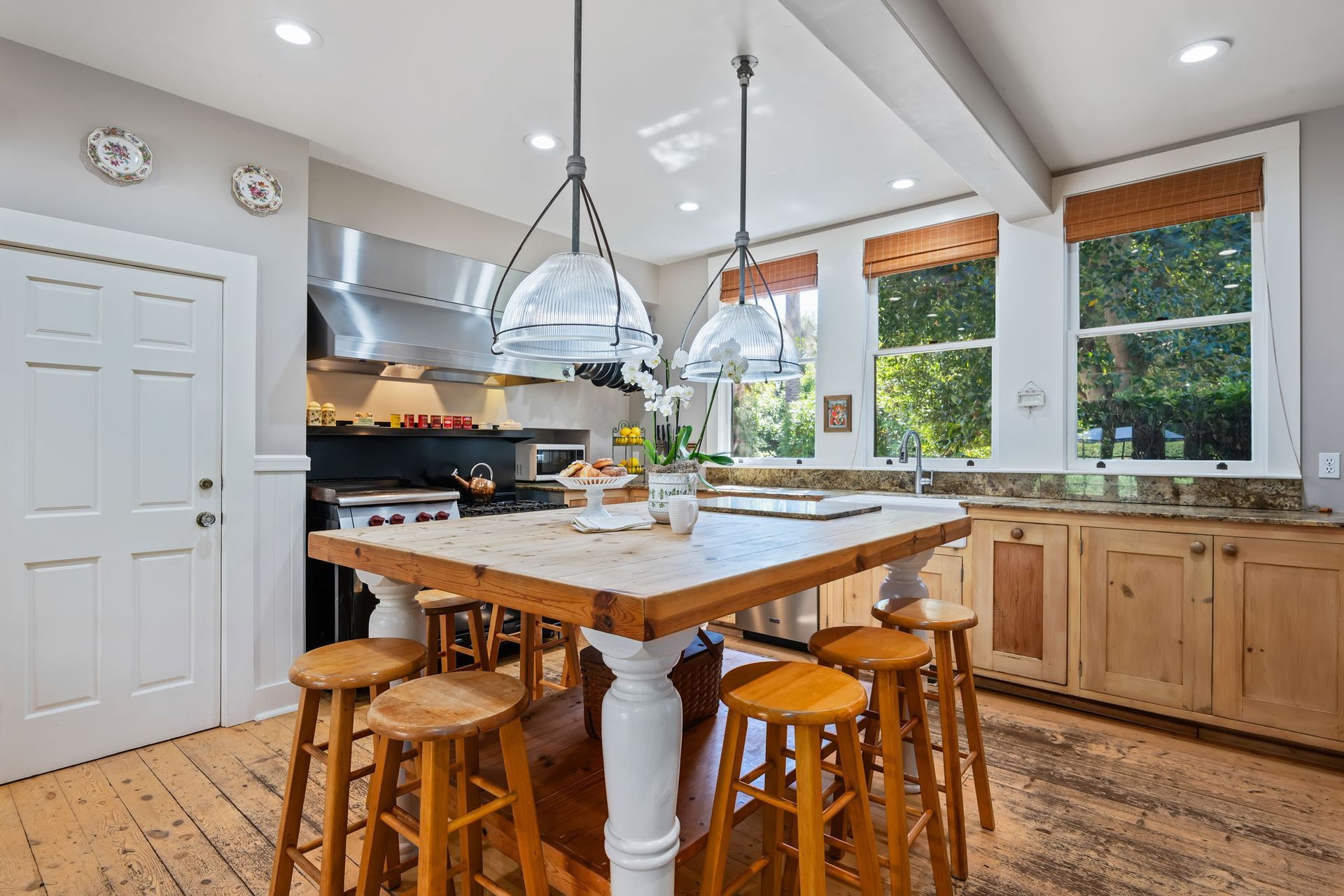 Bright kitchen with wooden island, stools, white cabinets, stainless hood, and large windows with orange shades