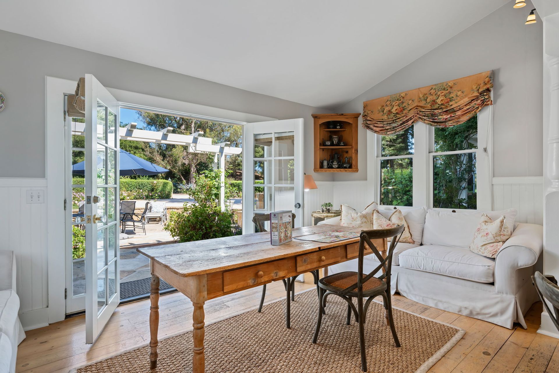 Bright coastal dining room with French doors open to a patio, wooden table, white sofa, and woven rug.