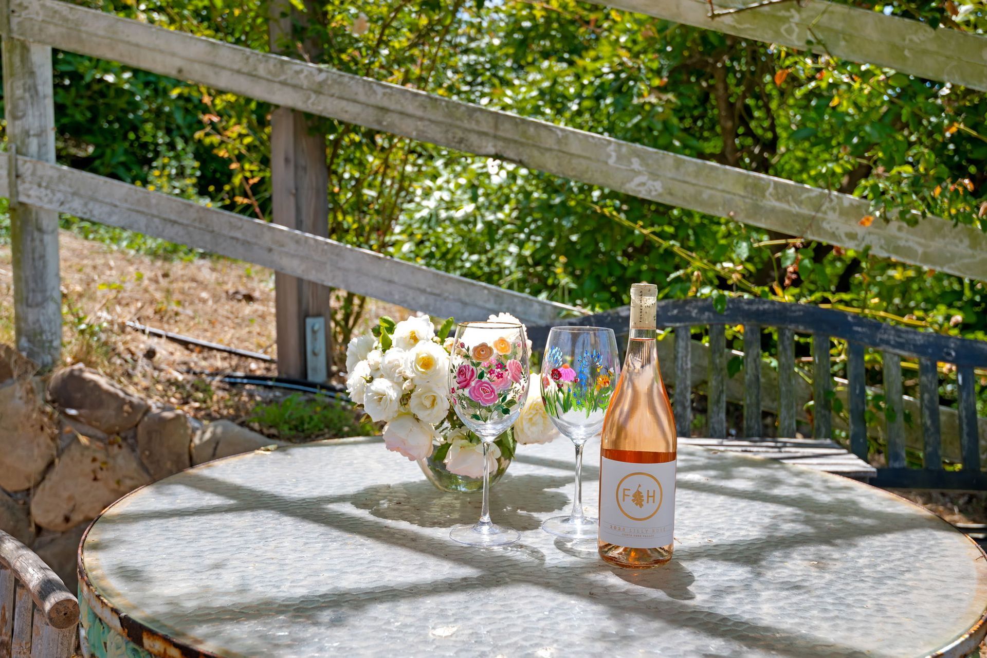 Round stone patio table with flowers and a bottle of rosé beside a wooden fence and greenery
